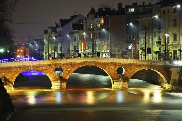 Eine beleuchtete Brücke in Sarajevo, Bosnien, bei Nacht. Die Lichter der Stadt spiegeln sich im Wasser.